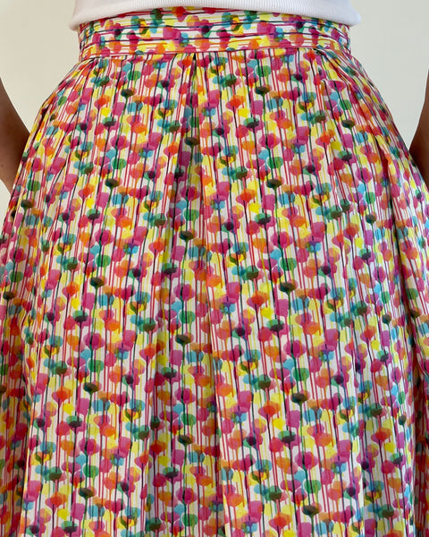 Backview, pleated waisted skirt. woman wearing fitted waistband skirt with pleats to mid calf. Has hands in pockets. bright splotchy colours of deep pink, blue, yellow green and orange on white cotton background