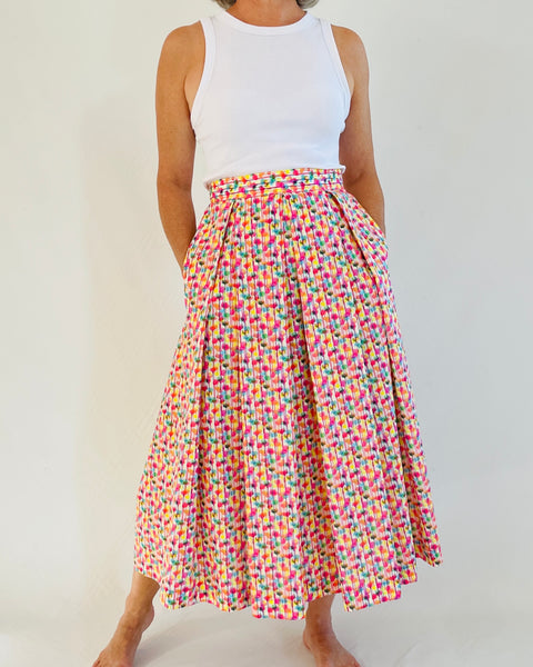 woman wearing fitted waistband skirt with pleats to mid calf. Has hands in pockets. bright splotchy colours of deep pink, blue, yellow green and orange on white cotton background