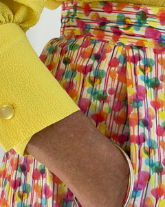 Closeup photo of woman in a bright yellow top. woman wearing fitted waistband skirt with pleats to mid calf. Has hands in pockets. bright splotchy colours of deep pink, blue, yellow green and orange on white cotton background