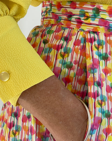 Closeup photo of woman in a bright yellow top. woman wearing fitted waistband skirt with pleats to mid calf. Has hands in pockets. bright splotchy colours of deep pink, blue, yellow green and orange on white cotton background