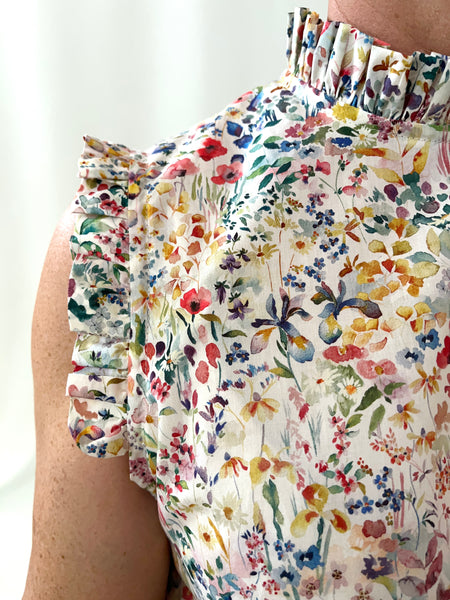 Woman wearing a floral cotton sleeveless dress against a white background. Dress has ruffles at neck and arms. Small floral print on cream-white background
