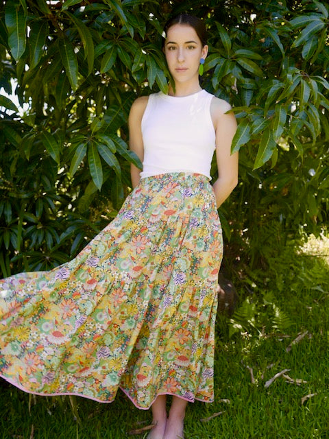 Woman standing in front of tree wearing a long calf length skirt, in green pinks and oranges floral design, 3 tiered with elastic waist