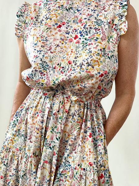 Woman wearing a floral cotton sleeveless dress against a white background. dress has ruffles at neck and arms. Elastic drawstring waist. two tier skirt that falls to lower shin. Small floral print on cream-white background