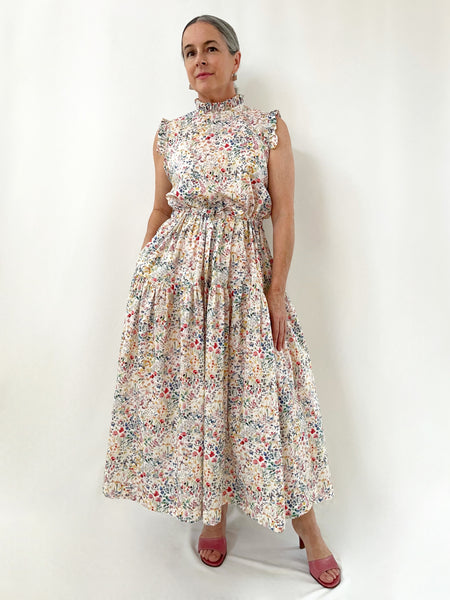 woman wearing a floral cotton sleeveless dress against a white background. dress has ruffles at neck and arms. Elastic drawstring waist. two tier skirt that falls to lower shin. Small floral print on cream-white background