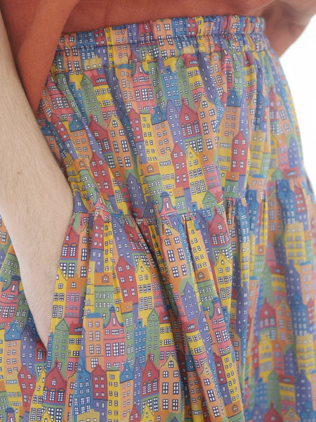 View of elastic waist band and side seam pockets. Woman wearing 3 tiered cotton printed skirt, knee length, elastic waisted with voluminous hem in mulitcoloured Copenhagen houses print. Colours are blue, rust red, mustard yellow, green.