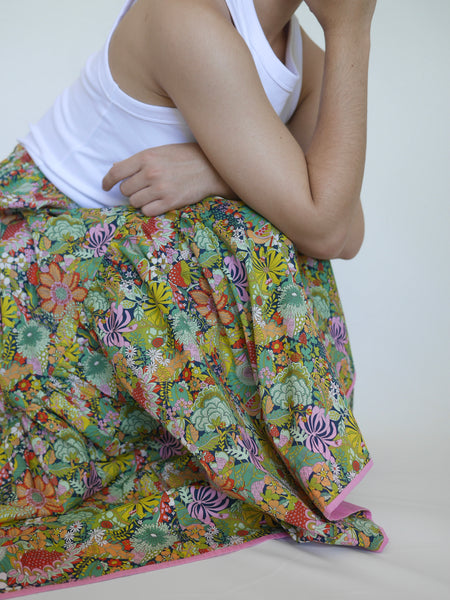 Woman wearing lower calf length 3 tiered skirt with elastic waist. With orange, pink, light green, navy, light orange, red detailed flowers printed on mid green background. Liberty print in light 100% cotton.. 1 cm candy pink edge on hem.