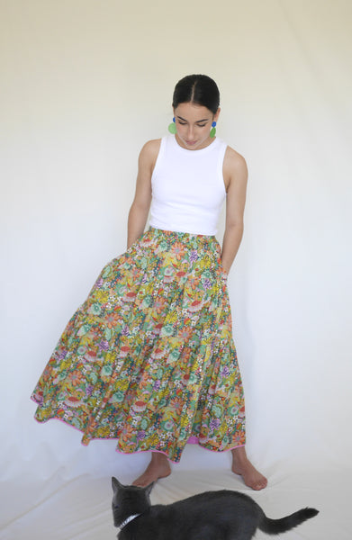 Woman talking to cat wearing whilst  lower calf length 3 tiered skirt with elastic waist. With orange, pink, light green, navy, light orange, red detailed flowers printed on mid green background. Liberty print in light 100% cotton.. 1 cm candy pink edge on hem.