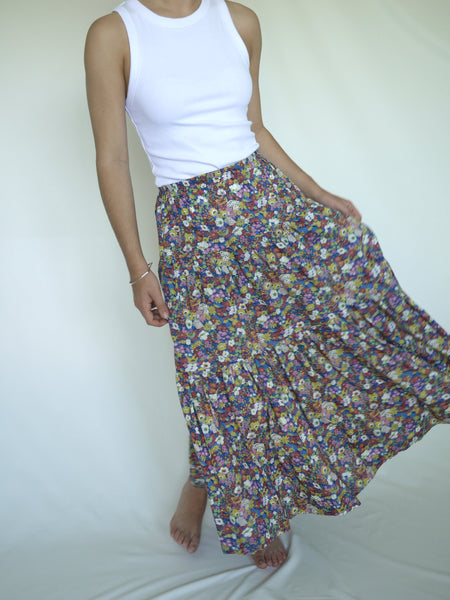 Woman wearing lower calf length 3 tiered skirt with elastic waist. White, yellow pink blue and green flower print on royal blue background. Liberty print in light 100% cotton.