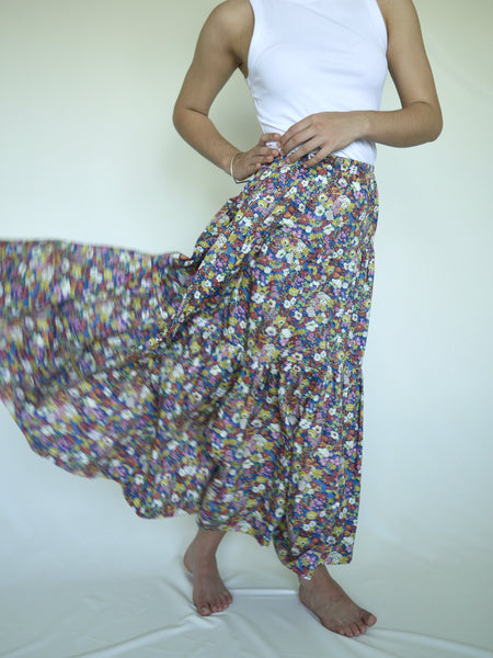 Woman  wearing lower calf length 3 tiered skirt with elastic waist. White, yellow pink blue and green flower print on royal blue background. Liberty print in light 100% cotton.