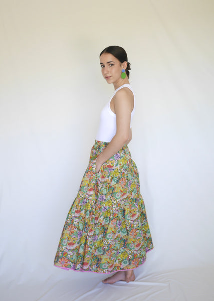 Woman wearing lower calf length 3 tiered skirt with elastic waist. With orange, pink, light green, navy, light orange, red detailed flowers printed on mid green background. Liberty print in light 100% cotton.. 1 cm candy pink edge on hem.