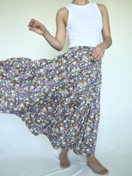 Woman wearing lower calf length 3 tiered skirt with elastic waist. White, yellow pink blue and green flower print on royal blue background. Liberty print in light 100% cotton.