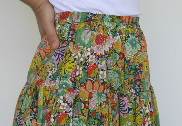 Woman wearing lower calf length 3 tiered skirt with elastic waist. With orange, pink, light green, navy, light orange, red detailed flowers printed on mid green background. Liberty print in light 100% cotton.. 1 cm candy pink edge on hem.