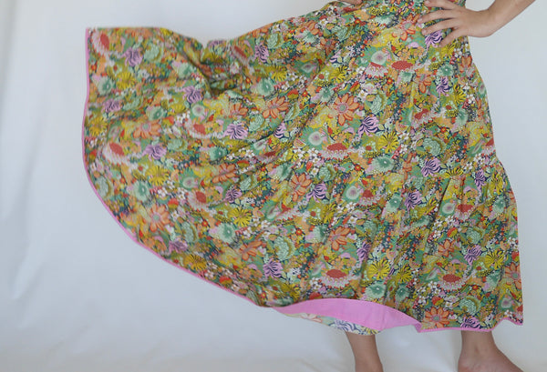 Woman swishing out her  lower calf length 3 tiered skirt with elastic waist. With orange, pink, light green, navy, light orange, red detailed flowers printed on mid green background. Liberty print in light 100% cotton.. 1 cm candy pink edge on hem.